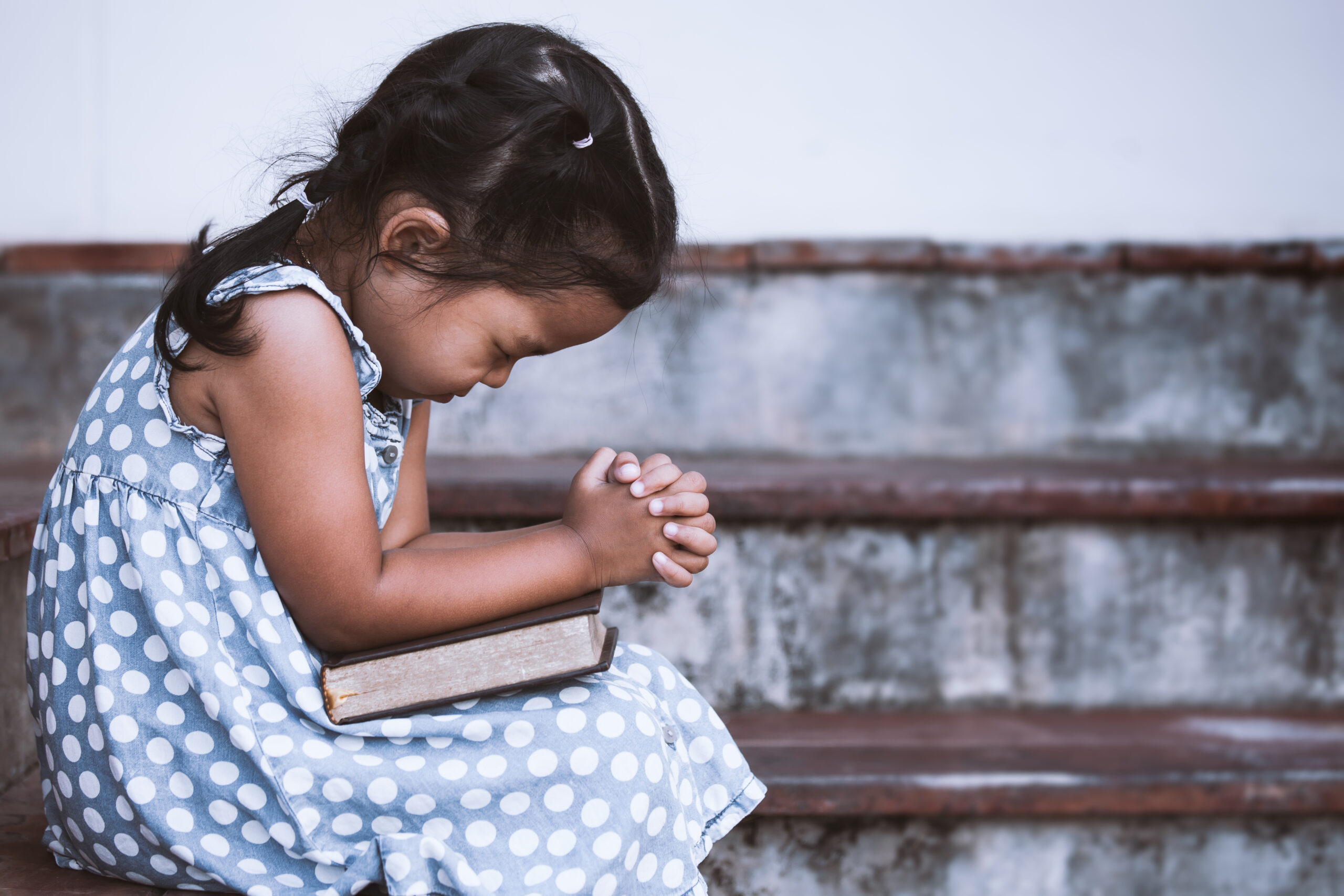 Cute asian little girl closed her eyes and folded her hand in prayer on a Holy Bible for faith concept in vintage color tone