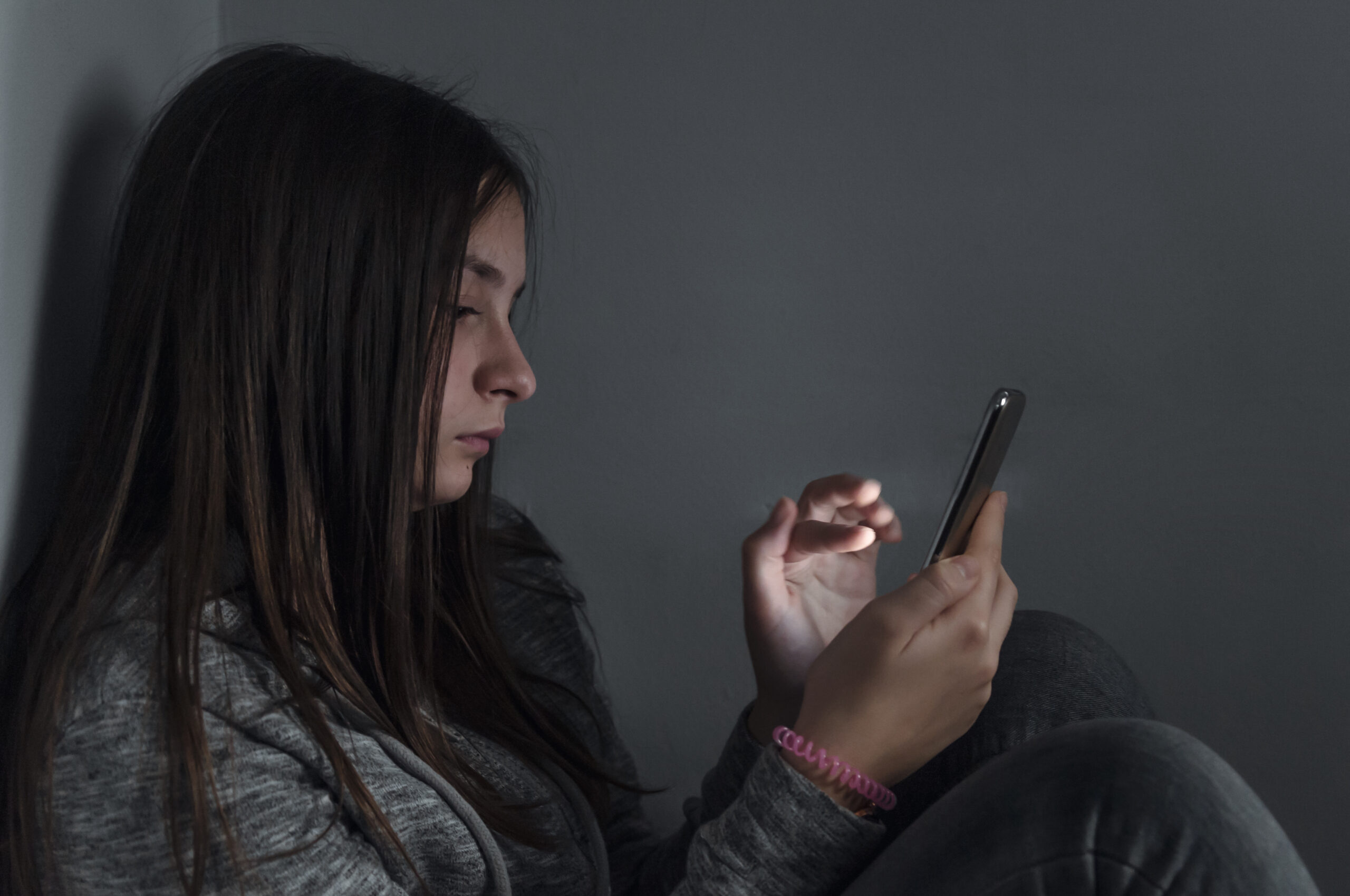 Teen girl excessively sitting at the phone at home. she is a victim of online social networks. Sad teen checking phone sitting on the floor in the living room at home with a dark background. Victim of online bullying Stalker social networks